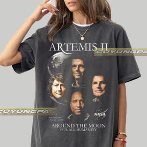 May include: Dark gray t-shirt featuring the text "ARTEMIS II" above portraits of four astronauts and a depiction of the moon. The shirt also includes the NASA logo and the phrase "AROUND THE MOON FOR ALL HUMANITY."