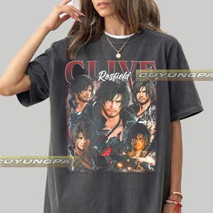 May include: Dark gray t-shirt featuring a graphic of a person with long hair. The text "CLIVE Rosfield" is printed in red and white. The shirt has short sleeves and a crew neck.