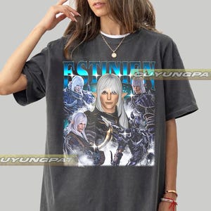May include: Dark gray t-shirt featuring a graphic of a character with white hair and the word "ESTINIEN" in blue. The design includes multiple figures in armor and a fantasy theme. The shirt has a vintage, worn look.