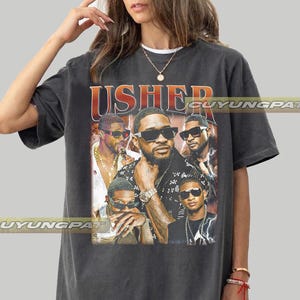 Może przedstawiać: Ciemnoszary t-shirt o kroju oversize z nadrukiem graficznym przedstawiającym artystę Ushera. Projekt zawiera napis USHER pogrubionymi pomarańczowymi literami nad kilkoma fotograficznymi portretami artysty w okularach przeciwsłonecznych i biżuterii.