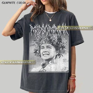 Può includere: Una t-shirt a maniche corte color grafite con una stampa grafica in bianco e nero di una persona che indossa una corona di fiori. Il testo MIDSOMMAR è stampato in un carattere stilizzato e frastagliato sopra il ritratto.