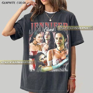 K&ouml;nnte beinhalten: Dunkelgraues T-Shirt mit einem Grafikdruck einer Frau und dem Text "JENNIFER Check" in Rot und Wei&szlig;. Die Grafik zeigt mehrere Bilder der Frau mit Vampir-Thema. Das Shirt hat eine lockere Passform.