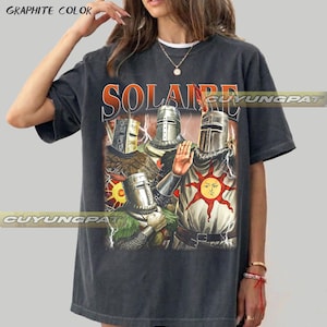 May include: A graphite-colored short-sleeve t-shirt featuring a graphic print of three armored knights in helmets. The central figure displays a red sun emblem on their chest. The text SOLAIRE is printed in large orange letters above the knights.