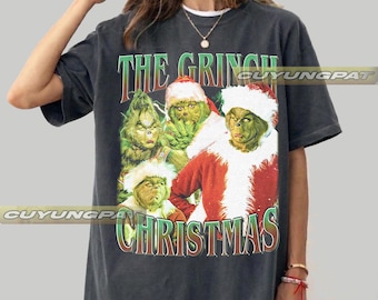 May include: A graphite-colored t-shirt featuring a graphic print of The Grinch in various poses, including one wearing a Santa suit. The text "THE GRINCH" appears in green at the top, and "CHRISTMAS" is printed in green at the bottom of the graphic.