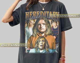 May include: A graphite-colored short-sleeve t-shirt featuring a graphic print from the movie Hereditary. The design includes the title text in yellow, along with portraits of three characters in various poses, set against a dark, vintage-style background.