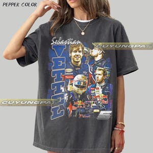 May include: A charcoal gray t-shirt featuring a graphic print of Formula 1 driver Sebastian Vettel. The design includes multiple images of the driver, his racing helmet, and the number 5, with the text Sebastian Vettel printed vertically.