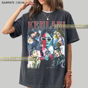 May include: A graphite-coloured short-sleeve t-shirt featuring a large graphic print of the artist Kehlani in various poses and outfits. The design includes the text KEHLANI in a bold red font at the top, with lightning motifs and vibrant colour accents.