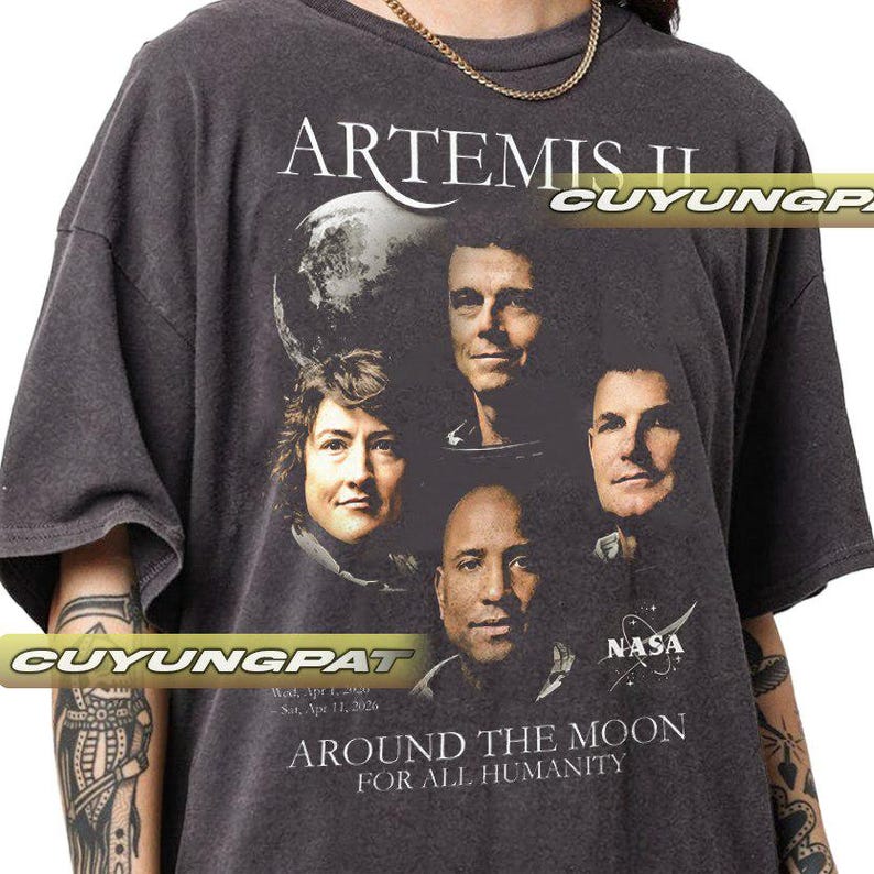 May include: Dark gray t-shirt featuring the text "ARTEMIS II" above a graphic of four people and a planet. Below the graphic, the shirt reads "AROUND THE MOON FOR ALL HUMANITY" and "NASA".
