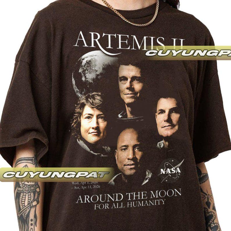 May include: Brown t-shirt featuring the text "ARTEMIS II" above a graphic of four people and a planet. Below the graphic, the text reads "AROUND THE MOON FOR ALL HUMANITY" and "NASA". The shirt has short sleeves.
