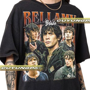 Puede incluir: Camiseta negra con un estampado gráfico de Bellamy Blake de la serie de televisión The 100. El gráfico presenta a Bellamy Blake con otros personajes de la serie. El texto "Bellamy Blake" está impreso en la camiseta.