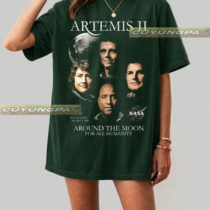 May include: Dark green t-shirt featuring the text "ARTEMIS II" and portraits of astronauts. The shirt also includes the NASA logo and the phrase "AROUND THE MOON FOR ALL HUMANITY". The shirt has short sleeves.