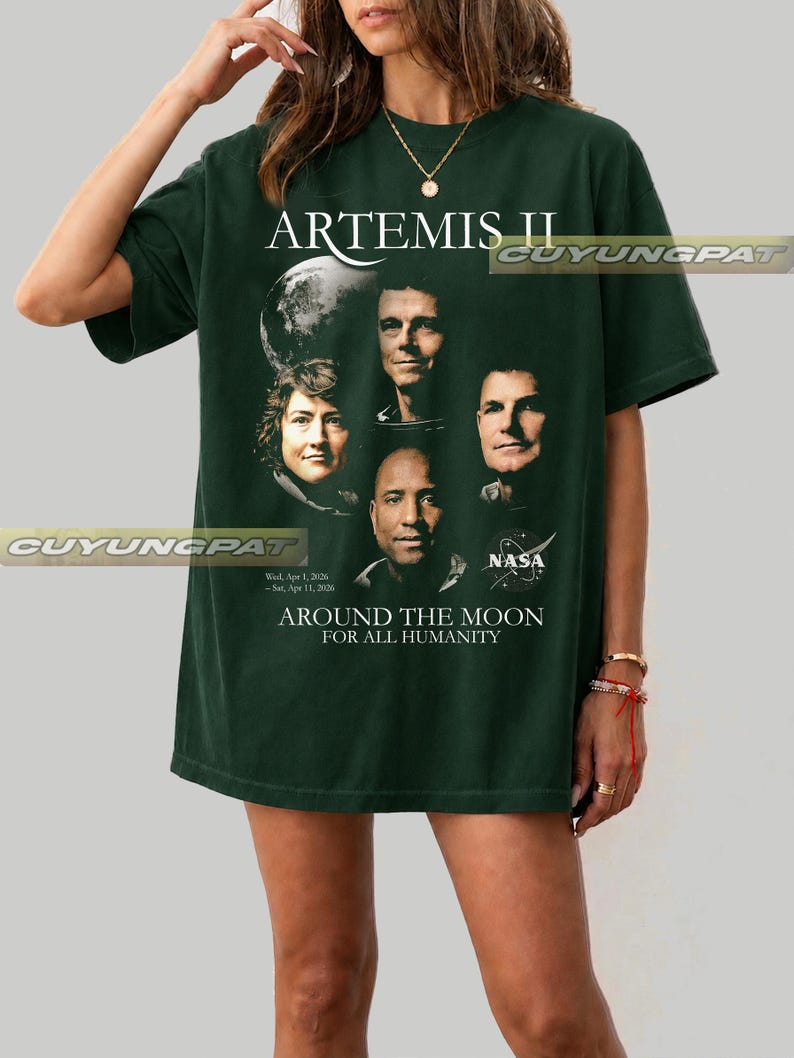May include: Dark green t-shirt featuring the text "ARTEMIS II" and portraits of astronauts. The shirt also includes the NASA logo and the phrase "AROUND THE MOON FOR ALL HUMANITY". The shirt has short sleeves.