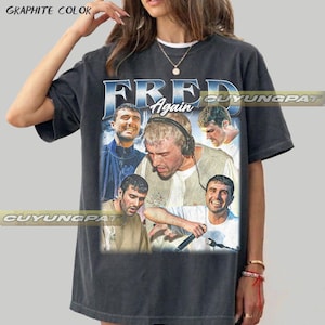 May include: Graphite-colored t-shirt featuring a collage of images of a person with the text "FRED Again" in blue and white. The shirt has a vintage, worn look.