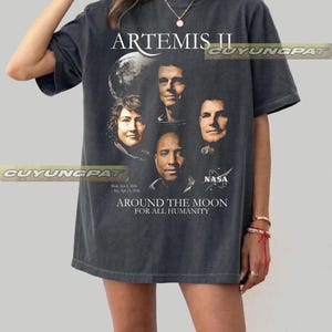 May include: Dark gray t-shirt featuring the text "ARTEMIS II" above a graphic of four astronauts and a planet. Below the image are the words "AROUND THE MOON FOR ALL HUMANITY" and the NASA logo.