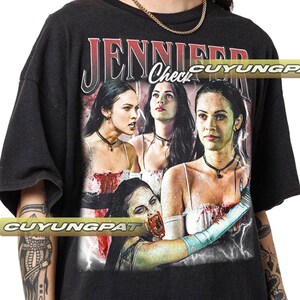 K&ouml;nnte beinhalten: Schwarzes T-Shirt mit einer Grafik einer Frau mit langen dunklen Haaren und dem Text "JENNIFER Check" in Rot und Wei&szlig;. Die Grafik hat einen Vintage-Look mit Blitz und einem Horror-Thema.