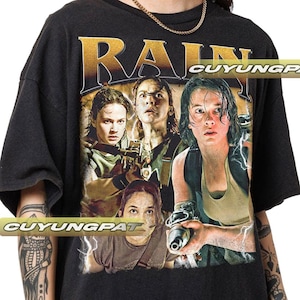May include: Black t-shirt featuring a graphic print of four characters from the film "Rain", with the word "RAIN" in gold lettering above them. The text "CUYUNGPAT" is printed at the bottom of the shirt.