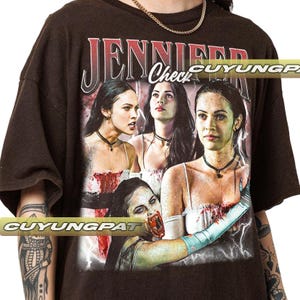 K&ouml;nnte beinhalten: Dunkelbraunes T-Shirt mit einer Grafik einer Frau und dem Text "JENNIFER Check". Die Grafik enth&auml;lt mehrere Bilder der Frau mit einem Horror-Thema. Das Shirt hat kurze &Auml;rmel und einen Rundhalsausschnitt.