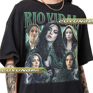 May include: Black t-shirt featuring a graphic of a woman with long dark hair and several other images of the same woman. The text "RIO VIDAL" is printed across the top in green letters. The shirt has a crew neck and short sleeves.
