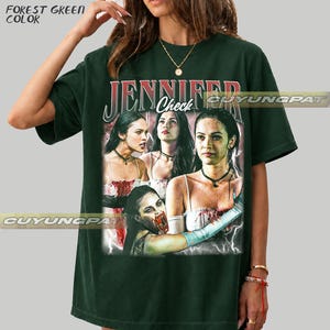 K&ouml;nnte beinhalten: Forstgr&uuml;nes T-Shirt mit einem Grafikdruck einer Frau und dem Text "JENNIFER Check". Die Grafik zeigt mehrere Bilder der Frau. Das T-Shirt hat eine dunkelgr&uuml;ne Farbe.
