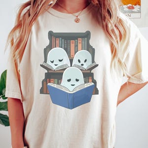 May include: Ivory-colored t-shirt featuring a whimsical illustration of three ghost characters reading books. The ghosts are light blue, and the books are various colors. The design is set against a backdrop of a bookshelf. The shirt is a casual, comfortable style.