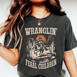 May include: A dark gray t-shirt with a vintage graphic of a cowboy riding a horse in a desert landscape. The text "WRANGLIN' FERAL CHILDREN" is printed on the shirt.