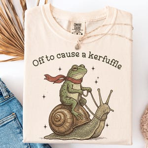 Op de afbeelding: Ivoor kleurig T-shirt met de tekst "Off to cause a kerfuffle". De afbeelding toont een kikker met een rode sjaal die op een slak rijdt. Het shirt ligt plat met een spijkerbroek en witte sneakers.