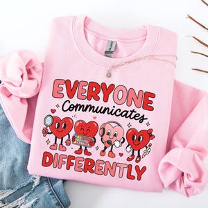 May include: A light pink sweatshirt with the text "EVERYONE Communicates DIFFERENTLY" in red and pink. The graphic features four cartoon hearts with different accessories. A pair of blue jeans is visible on the left.
