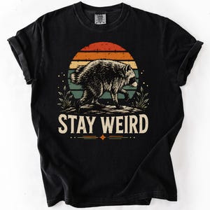 K&ouml;nnte beinhalten: Ein schwarzes T-Shirt mit einer Grafik eines Waschb&auml;ren, der vor einem Sonnenuntergangskreis mit orangefarbenen, gelben und blauen Streifen l&auml;uft. Unter dem Waschb&auml;ren steht der Text STAY WEIRD in einer fetten, hellen Schriftart.