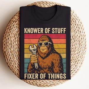 Puede incluir: Camiseta negra con un dise&ntilde;o retro. Presenta un personaje de Bigfoot con gafas de sol y una llave inglesa. El texto dice "KNOWER OF STUFF FIXER OF THINGS" en un estilo vintage con un fondo a rayas de colores.