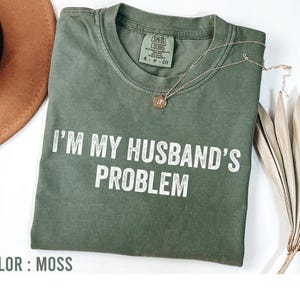 May include: A moss-colored t-shirt with the text "I'M MY HUSBAND'S PROBLEM" in white block letters. The shirt has a crew neck and short sleeves. A gold necklace with a pendant is draped on the shirt.