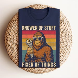 Puede incluir: Camiseta azul marino con un dise&ntilde;o retro. Presenta un personaje de Bigfoot con gafas de sol y una llave inglesa. El texto dice "KNOWER OF STUFF FIXER OF THINGS" con un fondo a rayas de colores.