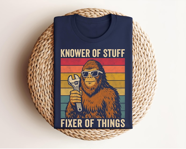 Puede incluir: Camiseta azul marino con un dise&ntilde;o retro. Presenta un personaje de Bigfoot con gafas de sol y una llave inglesa. El texto dice "KNOWER OF STUFF FIXER OF THINGS" con un fondo a rayas de colores.