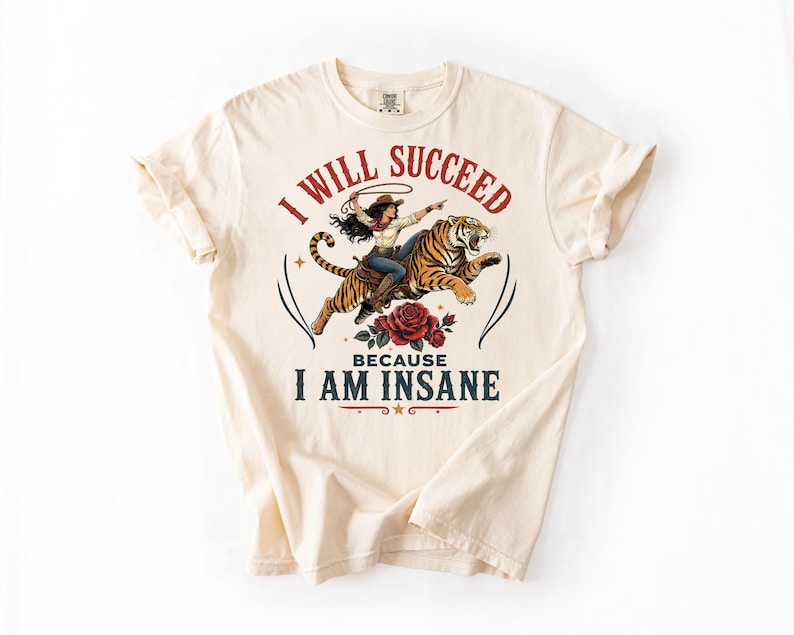 May include: A cream-colored t-shirt featuring a graphic of a person riding a tiger while holding a lasso. Below the tiger is a red rose. The text reads I WILL SUCCEED in red and BECAUSE I AM INSANE in dark blue, with small stars and decorative lines.