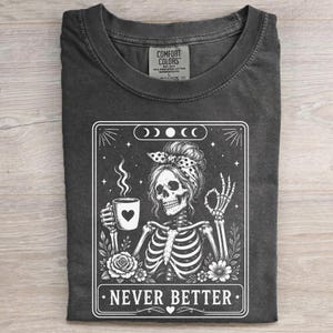 May include: A charcoal grey crew neck t-shirt featuring a white graphic print of a skeleton wearing a polka dot headscarf, holding a coffee mug with a heart, and making an okay hand gesture. The text "NEVER BETTER" is printed at the bottom.