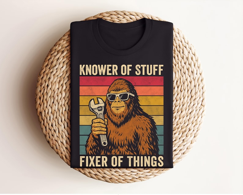 Puede incluir: Camiseta negra con un dise&ntilde;o retro. Presenta un personaje de Bigfoot con gafas de sol y una llave inglesa. El texto dice "KNOWER OF STUFF FIXER OF THINGS" en un estilo vintage con un fondo a rayas de colores.