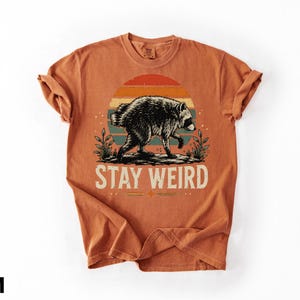 K&ouml;nnte beinhalten: Ein rostorangefarbenes Kurzarm-T-Shirt mit einer Grafik eines Waschb&auml;ren, der vor einem mehrfarbigen Sonnenuntergangskreis l&auml;uft. Unter dem Waschb&auml;ren ist der Text STAY WEIRD in einer hellen Schriftart gedruckt, flankiert von kleinen Pflanzen.