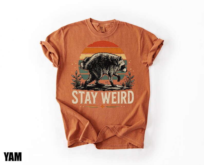 K&ouml;nnte beinhalten: Ein rostorangefarbenes Kurzarm-T-Shirt mit einer Grafik eines Waschb&auml;ren, der vor einem mehrfarbigen Sonnenuntergangskreis l&auml;uft. Unter dem Waschb&auml;ren ist der Text STAY WEIRD in einer hellen Schriftart gedruckt, flankiert von kleinen Pflanzen.