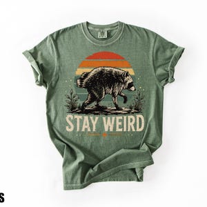 K&ouml;nnte beinhalten: Ein moosgr&uuml;nes Kurzarm-T-Shirt mit einer Grafik eines Waschb&auml;ren, der vor einem Sonnenuntergang mit orangefarbenen und blauen Streifen l&auml;uft. Unter dem Waschb&auml;ren ist der Text STAY WEIRD in einer fetten, hellen, serifenlosen Schrift gedruckt.