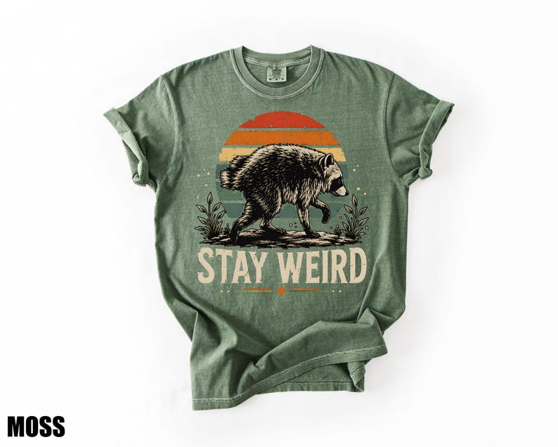 K&ouml;nnte beinhalten: Ein moosgr&uuml;nes Kurzarm-T-Shirt mit einer Grafik eines Waschb&auml;ren, der vor einem Sonnenuntergang mit orangefarbenen und blauen Streifen l&auml;uft. Unter dem Waschb&auml;ren ist der Text STAY WEIRD in einer fetten, hellen, serifenlosen Schrift gedruckt.