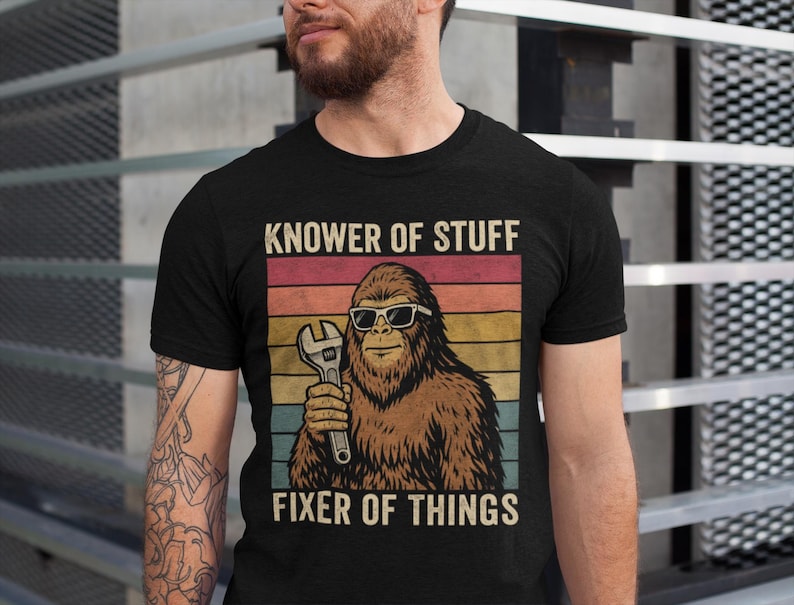 Puede incluir: Camiseta negra con un dise&ntilde;o retro que presenta un personaje de Bigfoot con gafas de sol y una llave inglesa. El texto "KNOWER OF STUFF" y "FIXER OF THINGS" est&aacute; impreso encima y debajo del gr&aacute;fico en un estilo vintage.