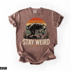 K&ouml;nnte beinhalten: Ein braunes Kurzarm-T-Shirt mit einer Grafik eines Waschb&auml;ren, der vor einem Sonnenuntergang mit orangefarbenen, gelben und t&uuml;rkisfarbenen Streifen l&auml;uft. Der Text "STAY WEIRD" ist in Wei&szlig; unter dem Waschb&auml;ren gedruckt. Das Wort "Espresso" ist zu sehen.