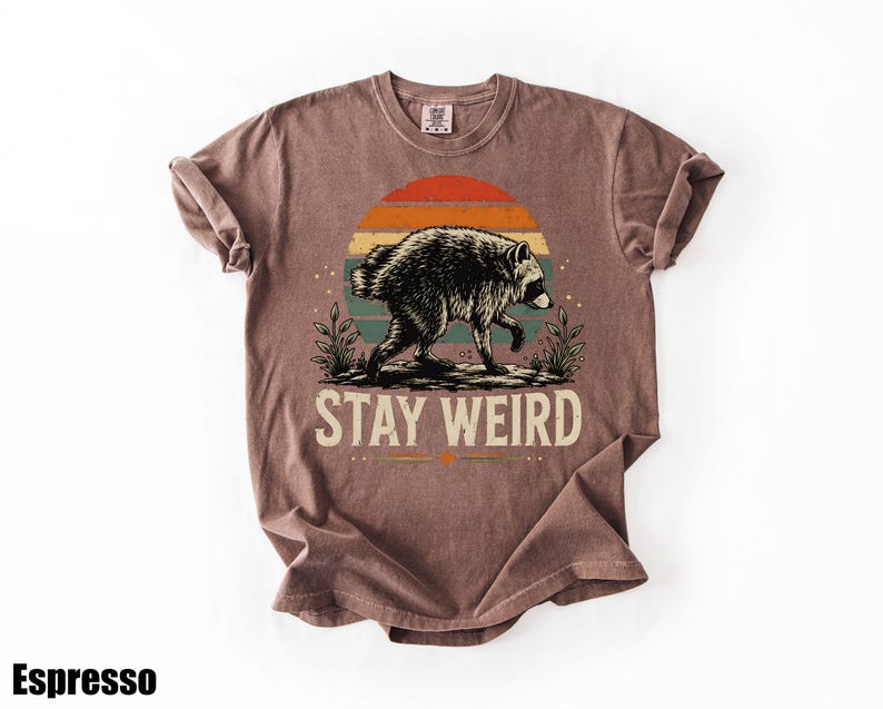 K&ouml;nnte beinhalten: Ein braunes Kurzarm-T-Shirt mit einer Grafik eines Waschb&auml;ren, der vor einem Sonnenuntergang mit orangefarbenen, gelben und t&uuml;rkisfarbenen Streifen l&auml;uft. Der Text "STAY WEIRD" ist in Wei&szlig; unter dem Waschb&auml;ren gedruckt. Das Wort "Espresso" ist zu sehen.