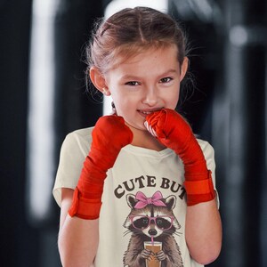 Pode incluir: Uma camiseta creme com um guaxinim de desenho animado usando &oacute;culos e la&ccedil;o rosa, segurando uma bebida. O texto "Cute But Feral" est&aacute; impresso abaixo. A pessoa est&aacute; usando ataduras de boxe vermelhas.