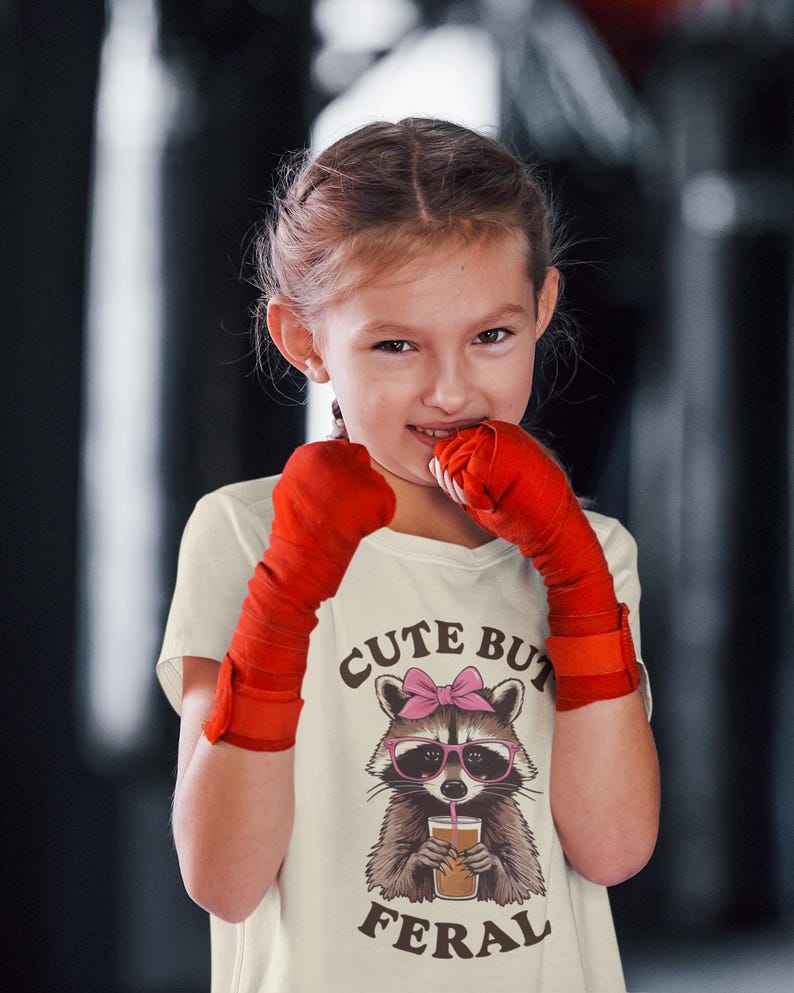 Pode incluir: Uma camiseta creme com um guaxinim de desenho animado usando &oacute;culos e la&ccedil;o rosa, segurando uma bebida. O texto "Cute But Feral" est&aacute; impresso abaixo. A pessoa est&aacute; usando ataduras de boxe vermelhas.