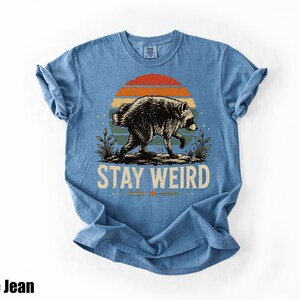 K&ouml;nnte beinhalten: Ein blaues T-Shirt mit einer Grafik eines Waschb&auml;ren, der vor einem Sonnenuntergang mit horizontalen Streifen in Orange, Gelb und Blaugr&uuml;n l&auml;uft. Unter dem Waschb&auml;ren ist der Text STAY WEIRD in einer fetten, hellen serifenlosen Schrift gedruckt.
