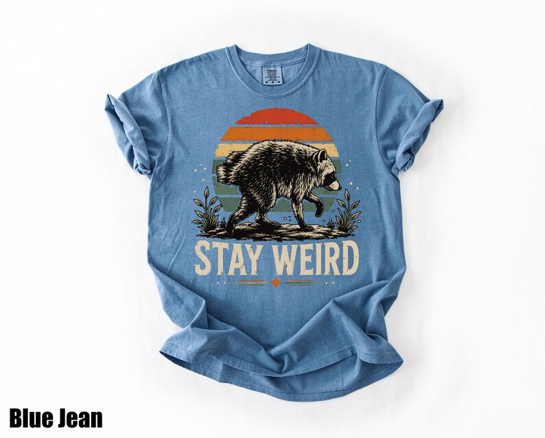 K&ouml;nnte beinhalten: Ein blaues T-Shirt mit einer Grafik eines Waschb&auml;ren, der vor einem Sonnenuntergang mit horizontalen Streifen in Orange, Gelb und Blaugr&uuml;n l&auml;uft. Unter dem Waschb&auml;ren ist der Text STAY WEIRD in einer fetten, hellen serifenlosen Schrift gedruckt.