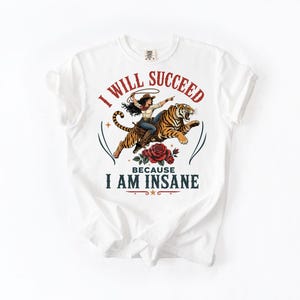 May include: A white short-sleeve t-shirt featuring a graphic of a cowgirl riding a tiger with a lasso. Below the tiger is a red rose. The text reads I WILL SUCCEED in red and BECAUSE I AM INSANE in dark blue, arranged in a circular arc.