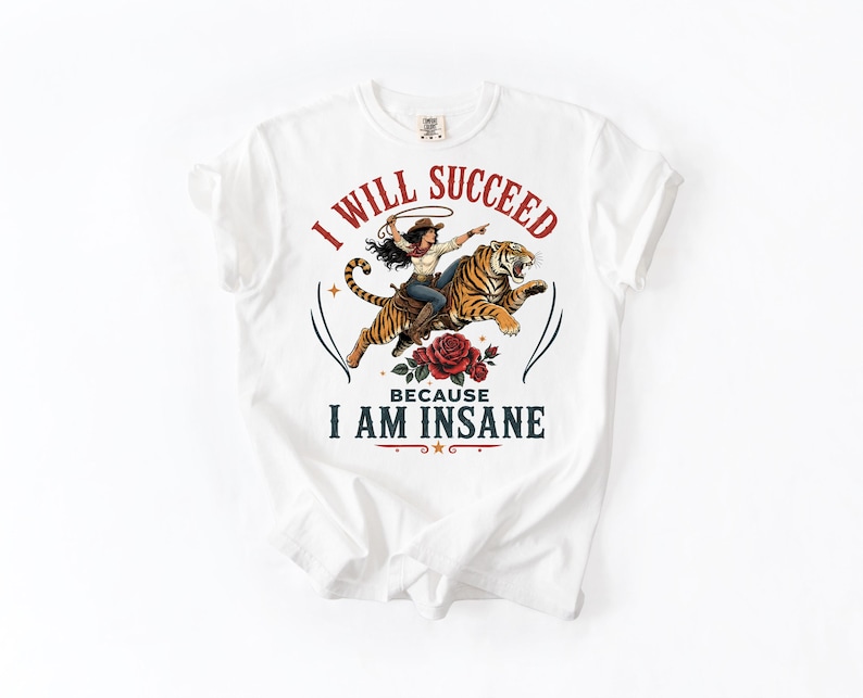 May include: A white short-sleeve t-shirt featuring a graphic of a cowgirl riding a tiger with a lasso. Below the tiger is a red rose. The text reads I WILL SUCCEED in red and BECAUSE I AM INSANE in dark blue, arranged in a circular arc.