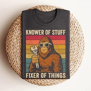 Puede incluir: Camiseta gris oscuro con un dise&ntilde;o retro. Presenta un personaje de Bigfoot con gafas de sol y una llave inglesa. El texto dice "KNOWER OF STUFF FIXER OF THINGS" en un estilo vintage.