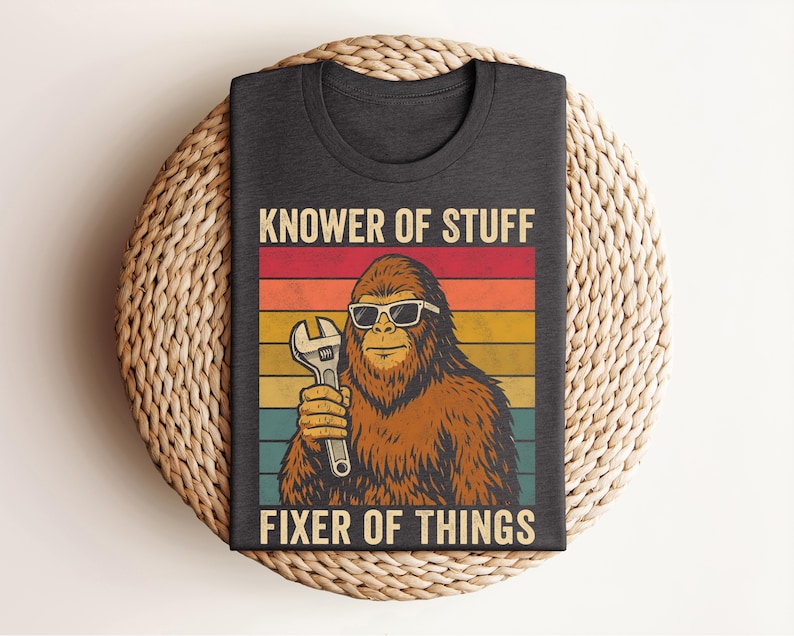 Puede incluir: Camiseta gris oscuro con un dise&ntilde;o retro. Presenta un personaje de Bigfoot con gafas de sol y una llave inglesa. El texto dice "KNOWER OF STUFF FIXER OF THINGS" en un estilo vintage.